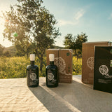 Two olive oil bottles and packaging on a table with a scenic background