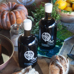 Two bottles of olive oil with a logo on a table with bread and vegetables