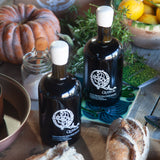 Two bottles of olive oil with a logo on a table with bread and vegetables