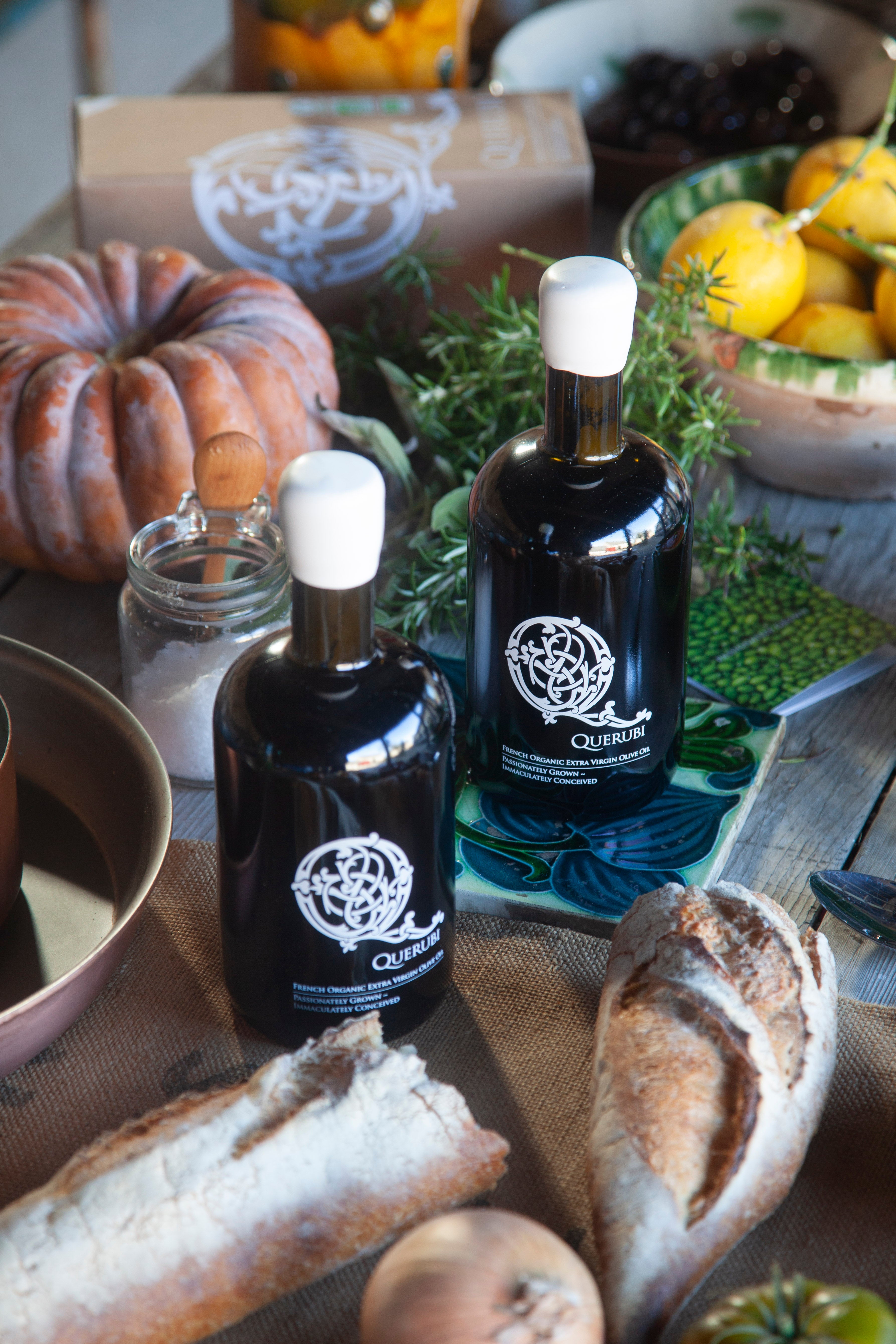 Two bottles of olive oil with a logo on a table with bread and vegetables